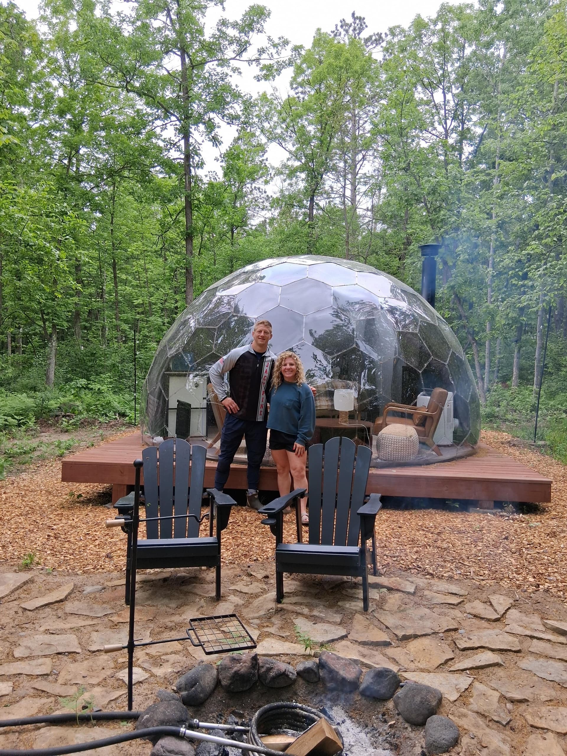 Guests inside a transparent Mirror Dome with Adirondack chairs and fire pit