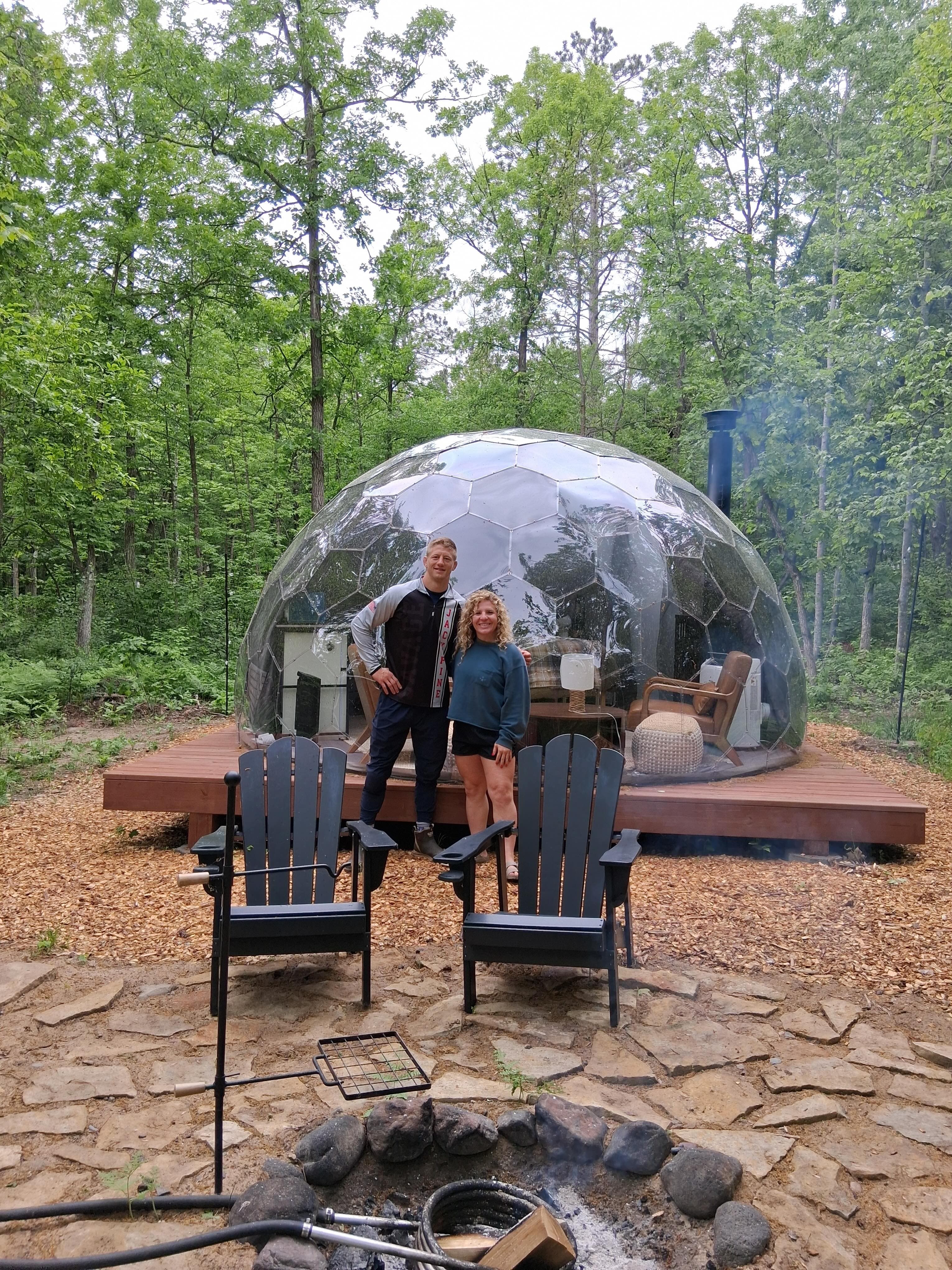 Two guests enjoying a transparent geodesic Mirror Dome in a forested setting